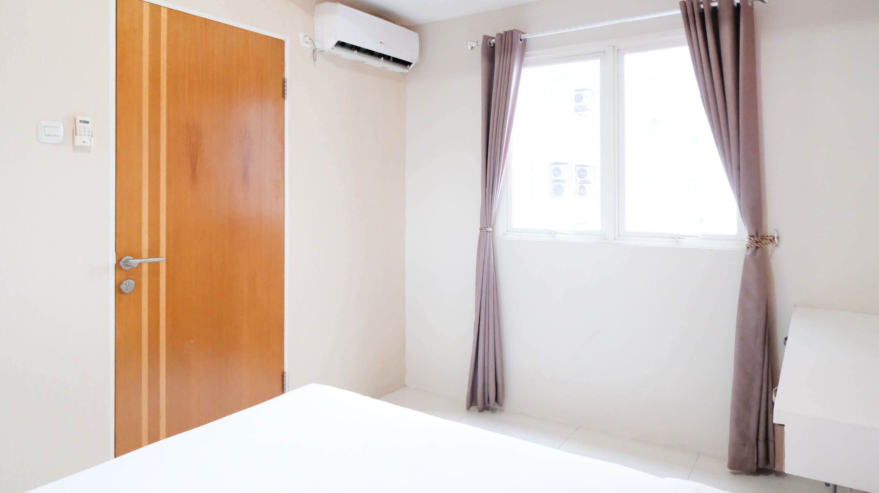 A room with a wooden door, an air conditioning unit, a window with curtains, and a white bed.