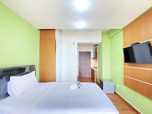 Quarto de hotel com uma cama, televisão fixada na parede e uma cozinha compacta ao fundo.