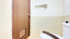 Une salle de bain avec une porte en bois, des carreaux blancs, des toilettes avec une serviette à proximité, et une étagère contenant des articles de toilette.