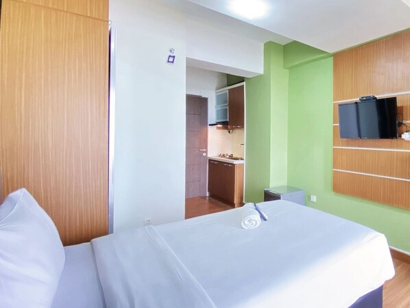 A hotel room with a bed, a TV mounted on the wall, and a kitchenette in the background.