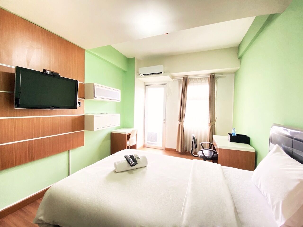 A hotel room with a bed, a TV mounted on the wall, a desk, a chair, and a window with curtains.