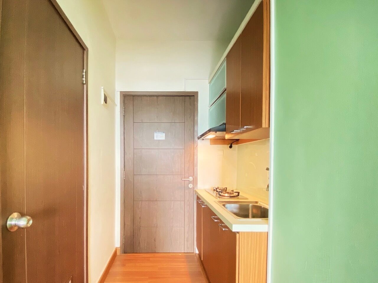 A narrow hallway leading to a closed door, with a kitchen area featuring cabinets and a sink on the right.