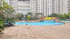 A swimming pool area with tiled flooring, surrounded by greenery and a building in the background. - Cozy Living And Tidy 2Br Green Bay Pluit Apartment (Jakarta)