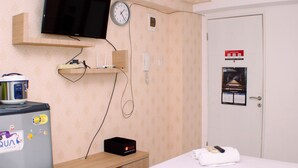 Une chambre d’hôtel avec un lit, un téléviseur fixé au mur, une horloge, une petite étagère, un réfrigérateur et une porte avec une pancarte.
