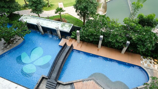 A large outdoor swimming pool with a waterfall feature, surrounded by lush greenery and a wooden deck. - Nice And Relax 1Br At Grand Sungkono Lagoon Apartment (Surabaya)