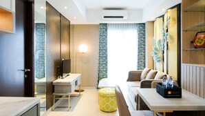A modern hotel room with a desk, a sofa, a small ottoman, and a dining table. There is a flat-screen TV on the desk, a wall-mounted air conditioner, and a window with curtains. - Nice And Relax 1Br At Grand Sungkono Lagoon Apartment (Surabaya)