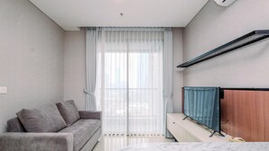 A modern hotel room with a grey sofa, a bed with a white headboard, a TV on a stand, and a large window with sheer curtains. - Nice And Strategic 1Br At Ciputra World 2 Apartment (Jakarta)