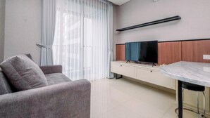 A modern living room with a sofa, a TV on a wooden cabinet, a dining table, and a large window with sheer curtains. - Nice And Strategic 1Br At Ciputra World 2 Apartment (Jakarta)