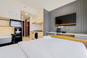 Quarto de hotel moderno com uma cama grande, televisão de tela plana na parede, uma pequena cozinha e um banheiro visível através de uma porta aberta.