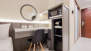 A compact hotel room with a bed, a desk with a chair, a microwave, a small trash bin, and a door. - Studio Room With Simply Look At Sky House Bsd Near Aeon (Pagedangan)