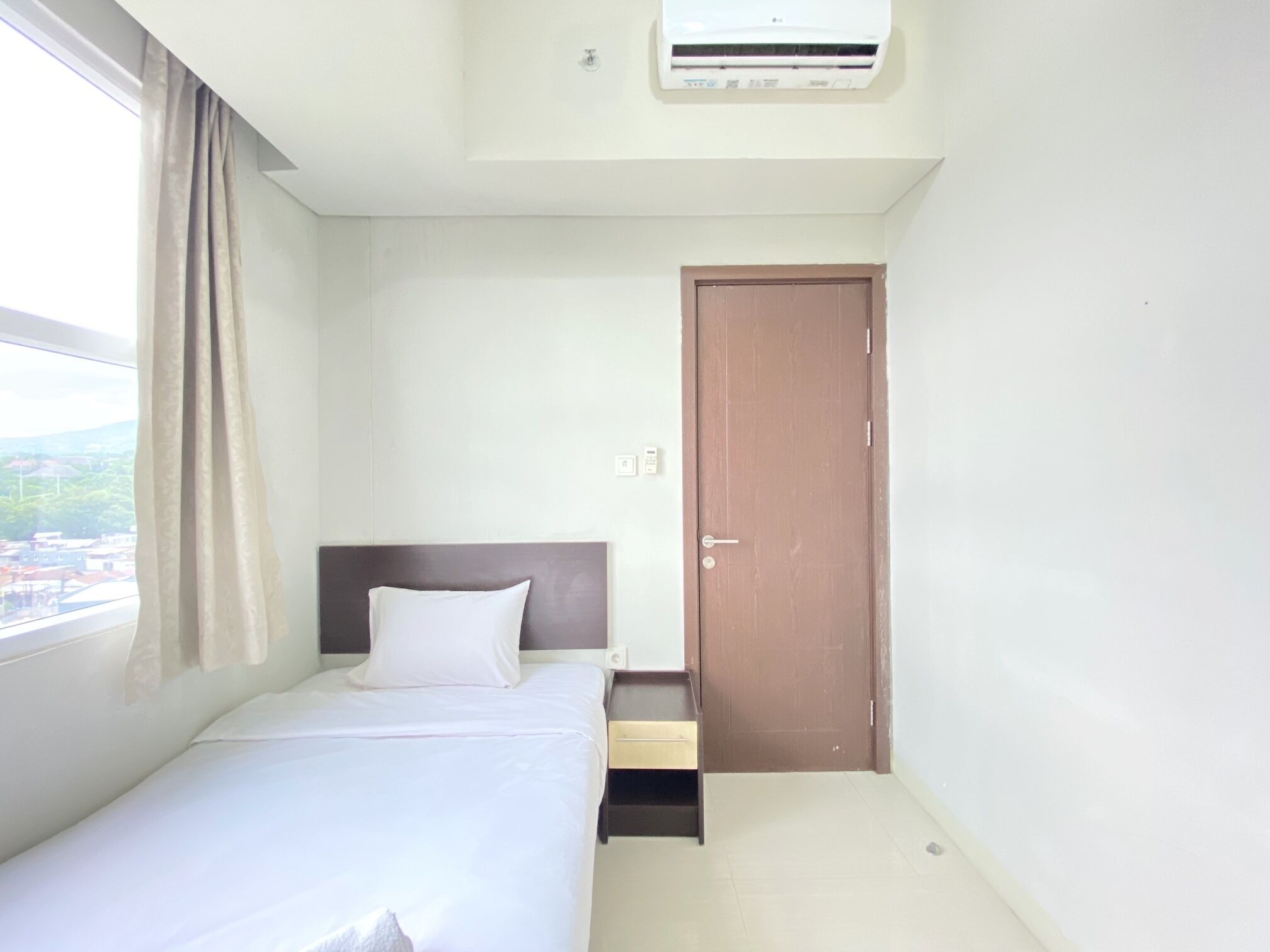 Una habitación de hotel con cama, ventana con cortinas, aire acondicionado, una puerta y una mesita.