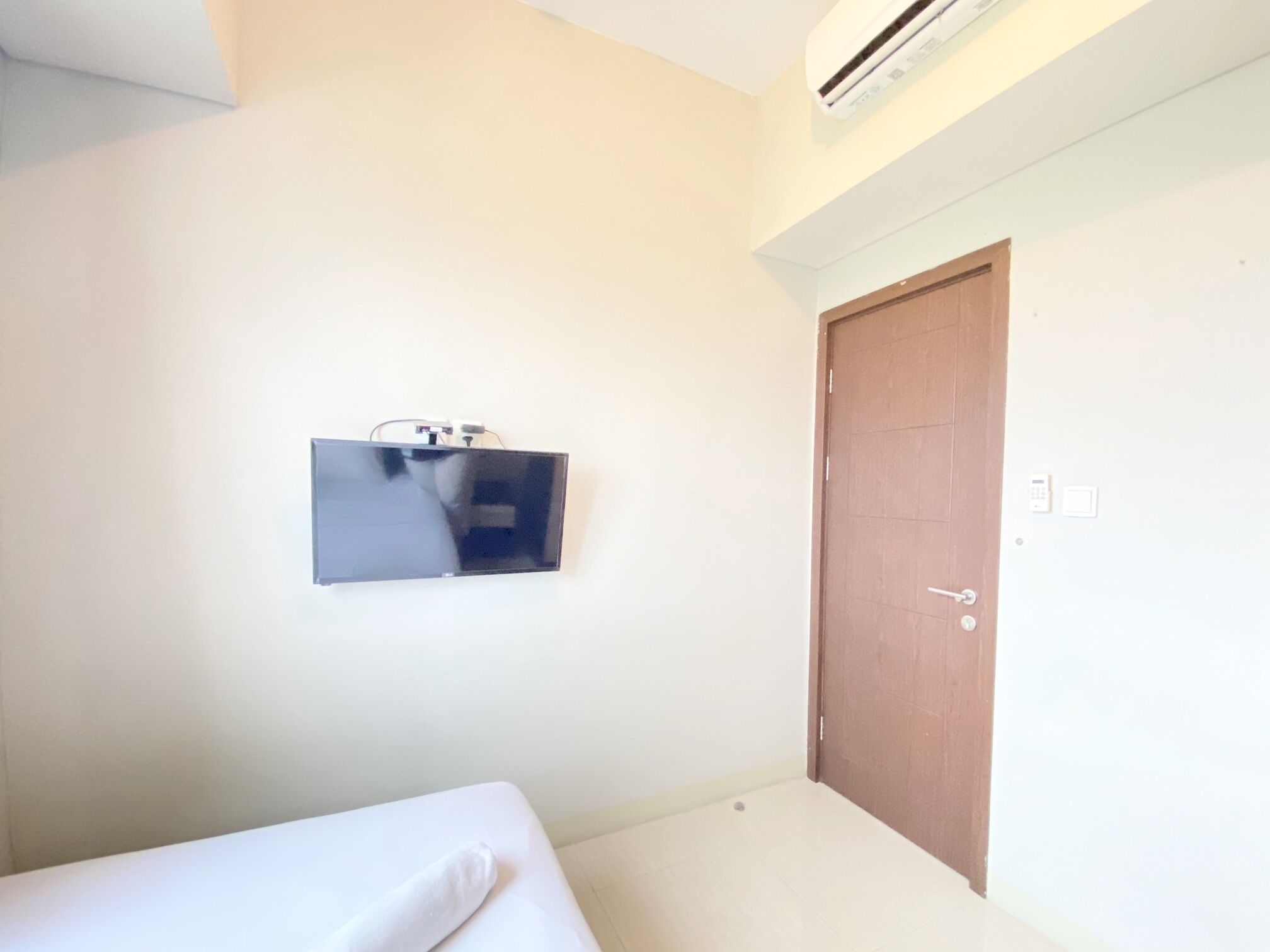 A room with a bed, a mounted television, a wooden door, and an air conditioning unit on the wall.