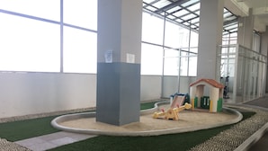 Un espace de jeux pour enfants avec un bac à sable et une petite structure en forme de maison, entouré de gazon synthétique et de galets.
