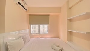 A hotel room with a bed, a window with blinds, a wall-mounted air conditioner, and a shelf unit. - Minimalist And Good Deal 2Br At Bassura City Apartment (Jakarta)
