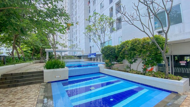 A swimming pool with a tiled deck, surrounded by greenery and a multi-story building in the background. - Good Deal And Minimalist 2Br At Bassura City Apartment (Jakarta)