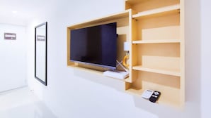 A modern room with a mounted TV, a wooden shelf unit, and a small table with a lamp and a remote control. - Cozy Studio At 9Th Floor Paltrow City Apartment (Semarang)