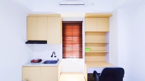 A compact kitchen with a built-in stove, sink, and a small countertop. There is a window with blinds, a chair, and a shelf unit with a green object on it. - Cozy Studio At 9Th Floor Paltrow City Apartment (Semarang)