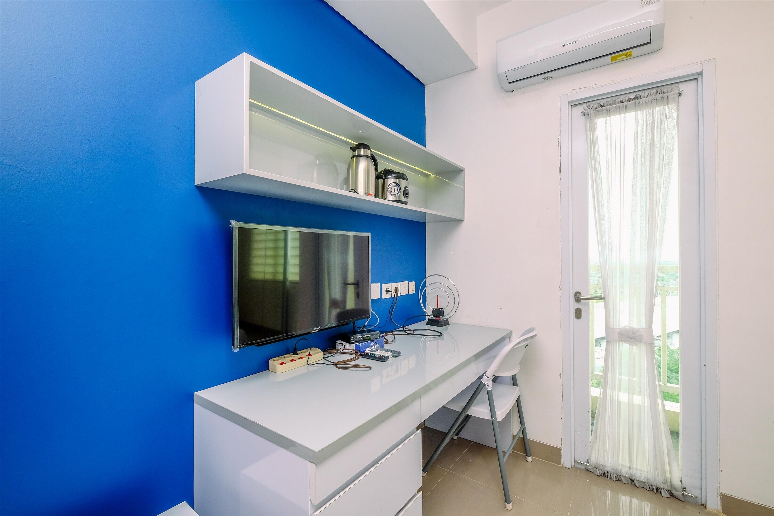 A modern room with a blue accent wall, a white desk with drawers, a mounted flat-screen TV, a shelf with kitchen appliances, and a door with a sheer curtain.