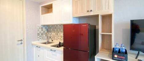 A compact kitchen with white cabinets, a red refrigerator, a tiled backsplash, and a flat-screen TV on a wooden shelf.