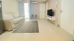 A living room with a sofa, a flat-screen TV, a rug, and a door. - Nice Comfortable 2Br Apartment At Bassura City (Jakarta)