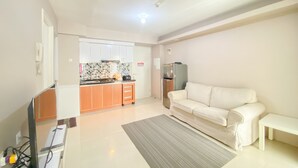A modern living room with a white sofa, a small table, and a kitchen area with orange cabinets and a tiled backsplash. - Nice Comfortable 2Br Apartment At Bassura City (Jakarta)