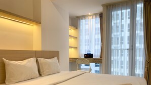 A hotel room with a large bed, two pillows, a desk with a chair, a window with curtains, and a minibar. - Cozy Studio At Tokyo Riverside Pik 2 Apartment (Teluknaga)