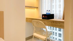 A hotel room with a wooden desk, a white chair, and a window with curtains. A black box labeled "Travelio" with two water bottles is on the desk. - Cozy Studio At Tokyo Riverside Pik 2 Apartment (Teluknaga)