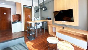 A modern living room with a flat-screen TV mounted on a wooden wall, a dining area with a long table and chairs, and a kitchen area with a brick backsplash and wooden cabinets. - Nice 2Br At Gateway Pasteur Apartment (Cimahi)