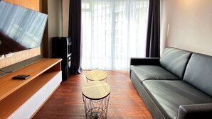 A modern living room with a flat-screen TV, a sofa, two small round tables, and a built-in speaker. - Nice 2Br At Gateway Pasteur Apartment (Cimahi)
