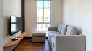 A modern living room with a flat-screen TV, a grey sofa, a wooden bench, and a window with a view of buildings. - Stylisth 1Br At Tokyo Riverside Pik 2 Apartment (Teluknaga)
