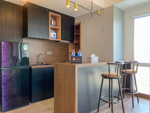 A modern kitchen with a refrigerator, wooden cabinets, a built-in island with bar stools, and a window providing natural light. - Stylisth 1Br At Tokyo Riverside Pik 2 Apartment (Teluknaga)