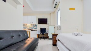 Ett hotellrum med en svart lädersoffa, en vit säng med hoprullade handdukar, ett skrivbord med en stol, en tv och ett litet kök i bakgrunden.