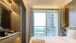 A hotel room with a bed, a TV, a minibar, and a view of a cityscape through the window.