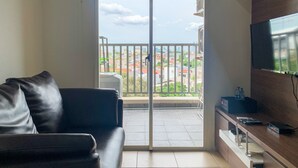 A living room with a sofa, a TV, and a balcony with a city view. - Warm And Homey 1Br At Belmont Residence Puri Apartment (Jakarta)