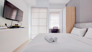 A modern hotel room with a large bed, a flat-screen TV mounted on the wall, a desk with a computer, and a window with blinds. - Enjoy Living And Warm Studio At Serpong Garden Apartment (Cisauk)