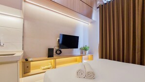 A modern hotel room with a flat-screen TV mounted on the wall, a wooden bedside table with a plant, and neatly rolled towels on the bed. - Scenic Designed And Comfy Studio At Sky House Bsd Apartment (Pagedangan)