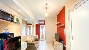 A modern living room with a sofa, TV, and a desk with a computer. There are two windows with curtains, an air conditioning unit, and a door with a phone on the wall. - Good Deal And Well Furnished 2Br At Springlake Summarecon Bekasi Apartment (Bekasi)