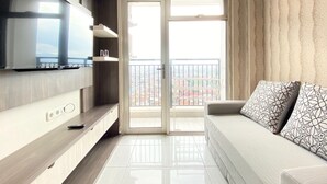 A modern living room with a flat-screen TV, a sofa with patterned cushions, and a sliding glass door leading to a balcony with a city view. - Chic And Cozy 2Br At Mekarwangi Square Cibaduyut Apartment (Bandung)