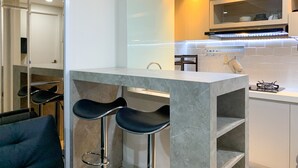 A modern kitchen with a marble countertop, a gas stove, and a built-in oven. There are two black bar stools and a chair. - Comfort And Enjoy Living 2Br At Daan Mogot City Apartment (Jakarta)