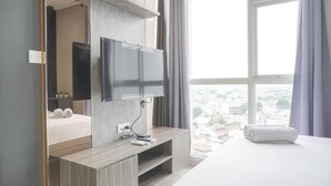 Una habitación de hotel moderna con un televisor grande montado en la pared, una cama con edredón blanco, una mesa consola de madera y vistas a la ciudad a través de amplios ventanales.