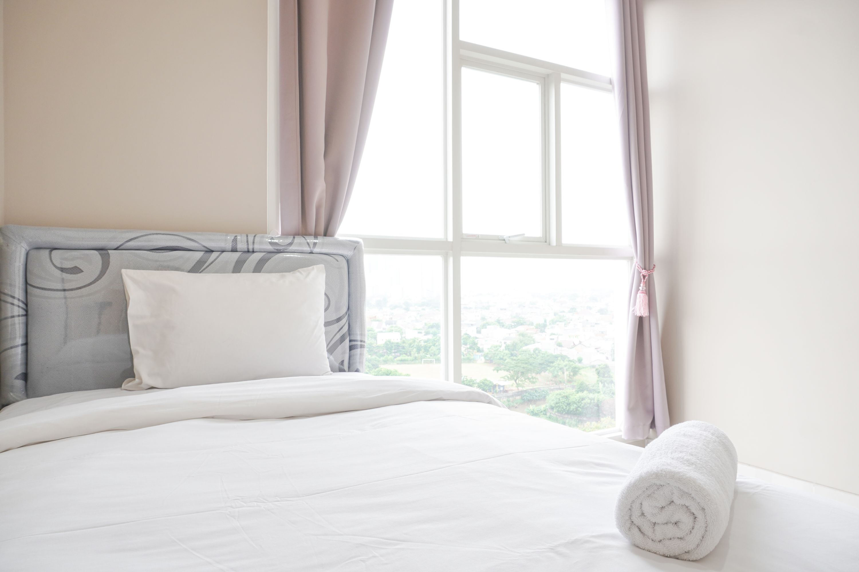 A neatly made bed with a white comforter and pillows, a rolled white towel, and a view of greenery through a window with curtains.