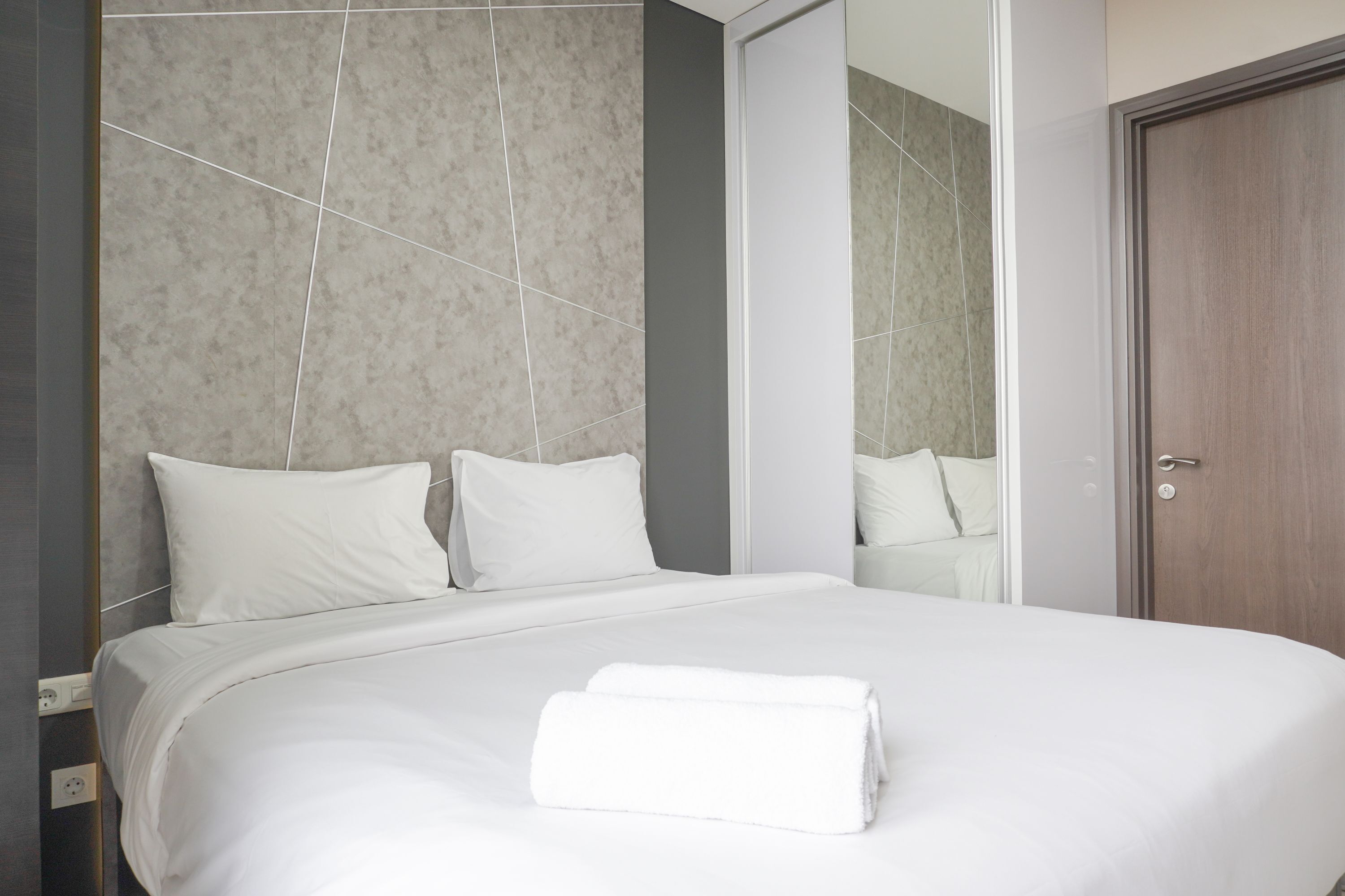 A neatly made bed with white linens and a folded towel, a wooden door, and a mirror reflecting part of the room.
