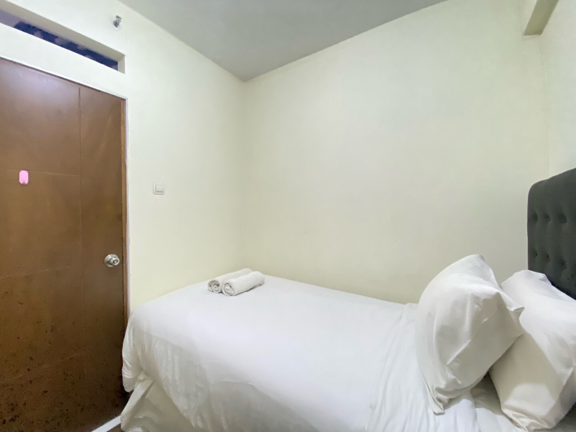 A small, single-bed room with a wooden door, white walls, and a grey headboard.