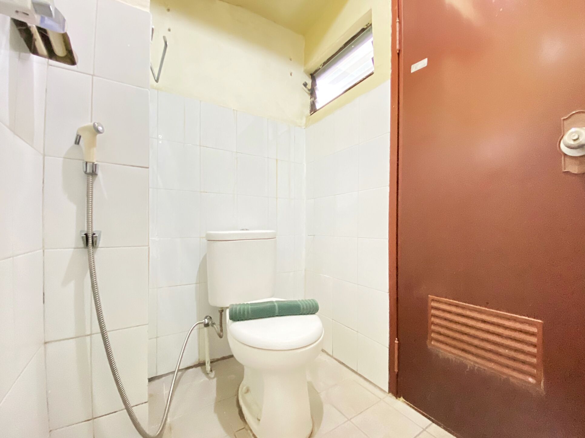 A bathroom with white tiled walls, a white toilet, a shower with a hose, and a brown door.