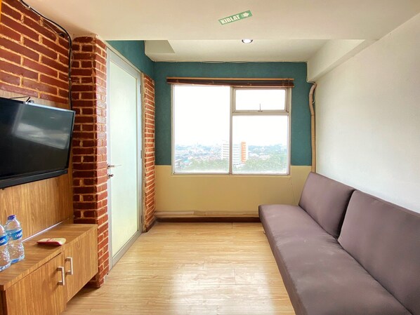 A room with a television on a wooden cabinet, a sofa, a window with a city view, and a brick wall. - Spacious And Unique 1Br At Jarrdin Cihampelas Apartment (Bandung)
