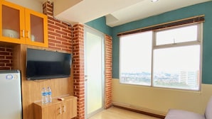 A room with a TV mounted on a brick wall, a wooden cabinet below, a window with a city view, and a grey sofa. - Spacious And Unique 1Br At Jarrdin Cihampelas Apartment (Bandung)