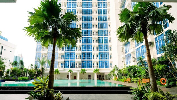 A hotel with a pool surrounded by palm trees and a modern building with blue windows in the background. - Nice And Strategic Studio Apartment At Capitol Park Residence (Jakarta)