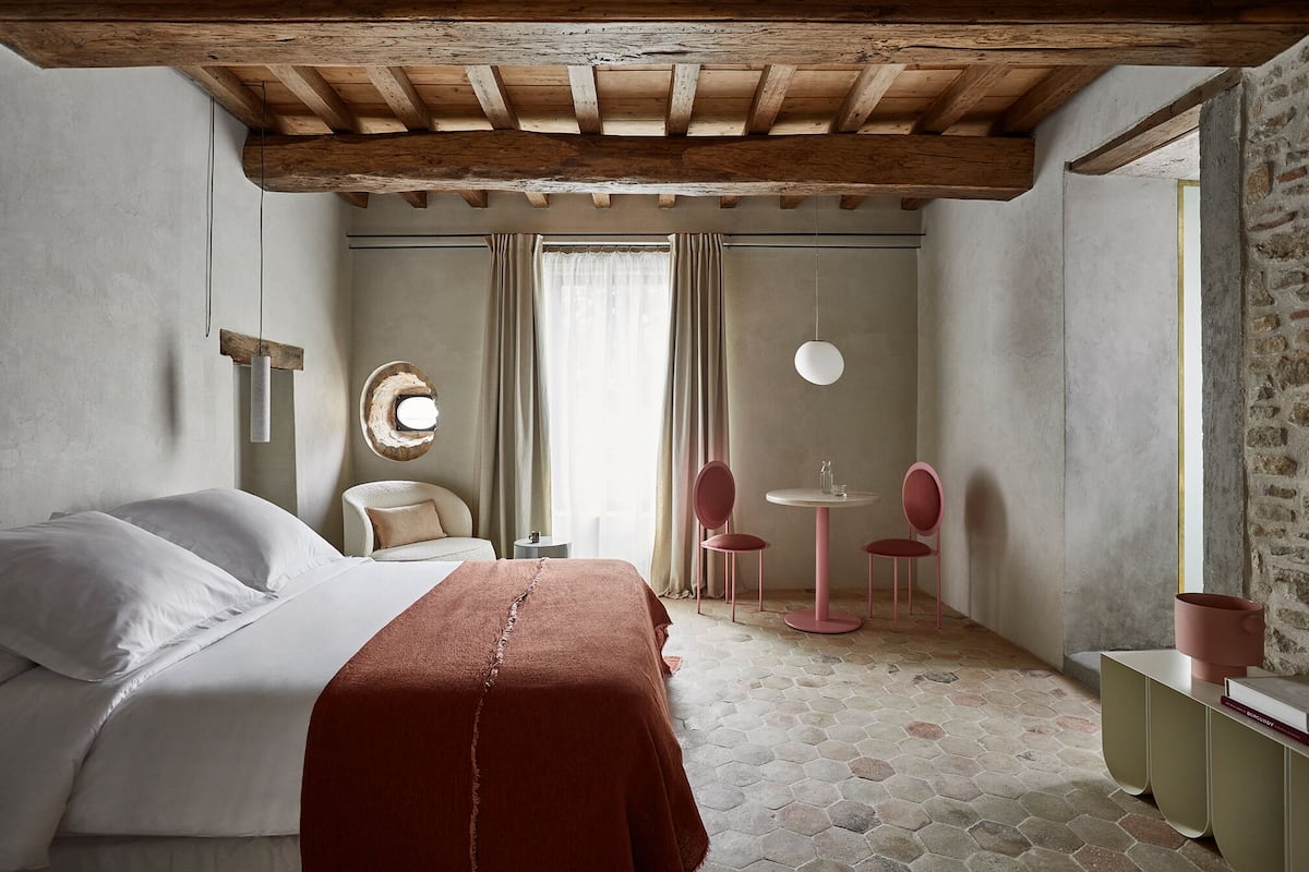 A bedroom with a large bed, a small table with chairs, a round mirror, and a window with curtains.