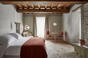 Een slaapkamer met een groot bed, een klein tafeltje met stoelen, een ronde spiegel en een raam met gordijnen.