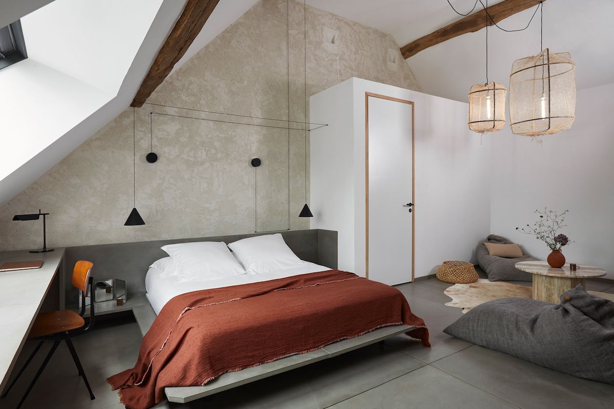 A modern bedroom with a sloped ceiling, a bed with a red blanket, a desk with a chair, and a small table with a vase.